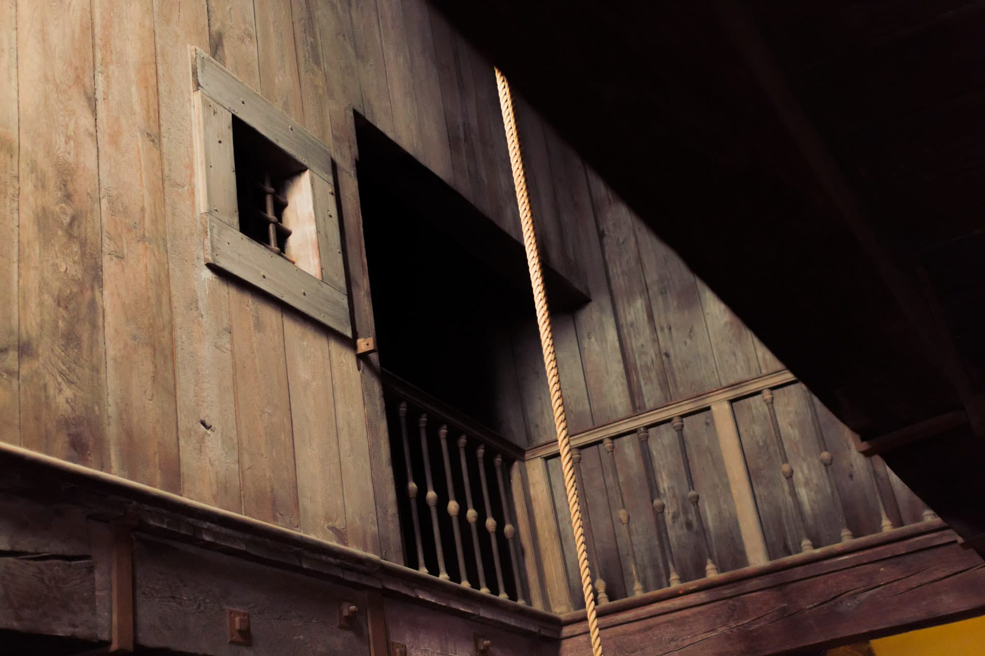 A wooden wall with a barred window and open balcony inside a concealed chamber, with a rope hanging nearby.