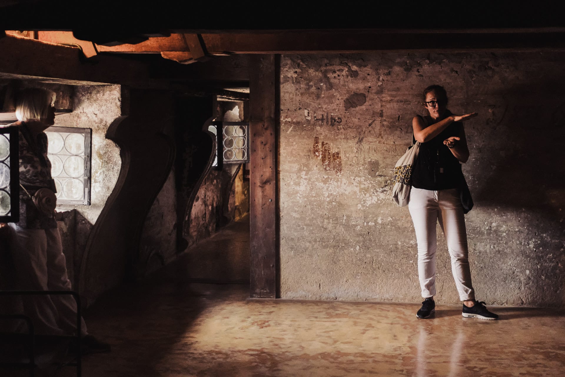 A guide explains something in a low-lit, rustic room with carved windows and worn stone walls inside doge's palace.