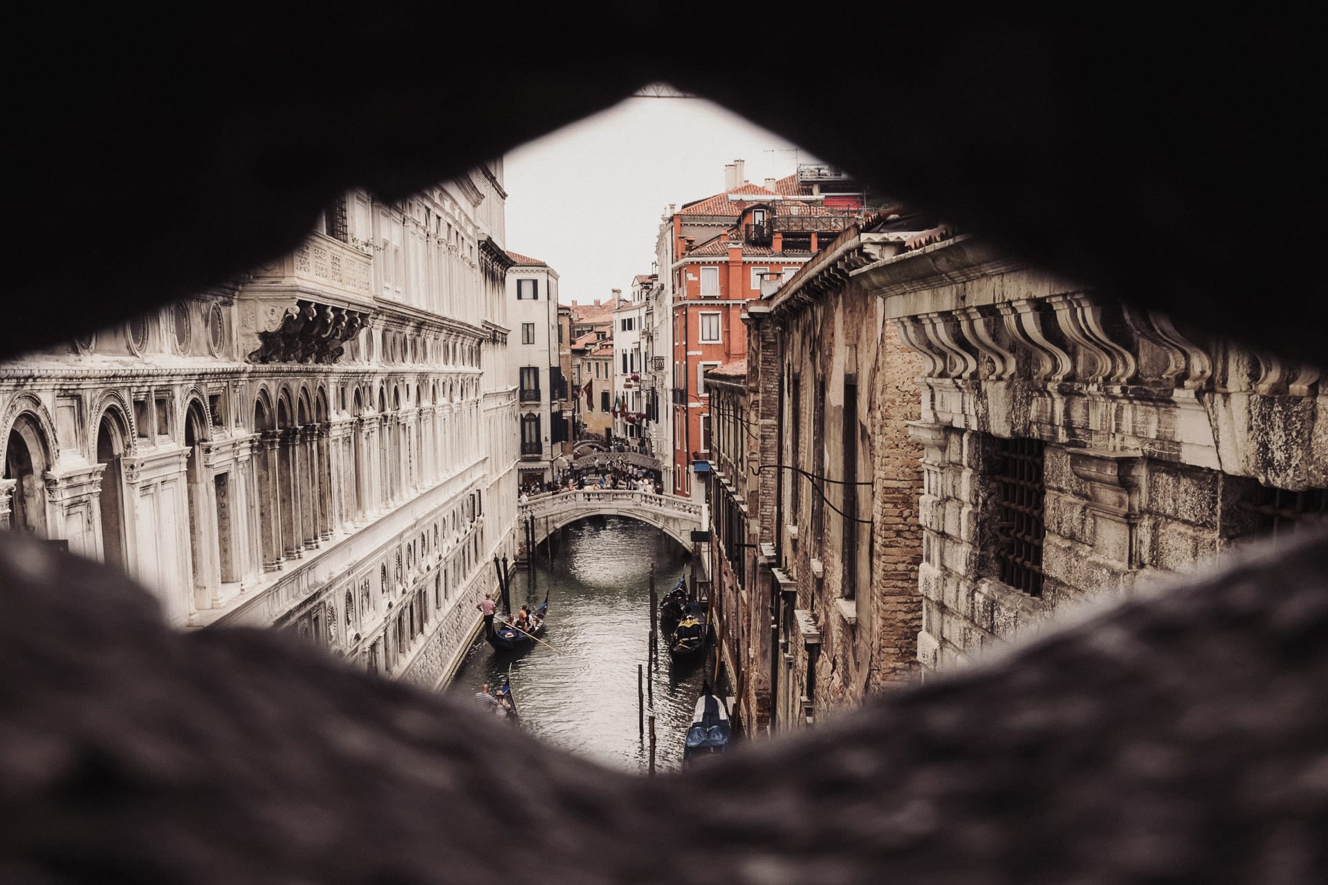 The view of a gondola ride in Venice, from one of the bridges above. Looking through a diamond shaped hole in the bridge you can see the intricate white stone architecture of the building on the left, contrasted against red and darker stone buildings on the right, and a bridge in the distance