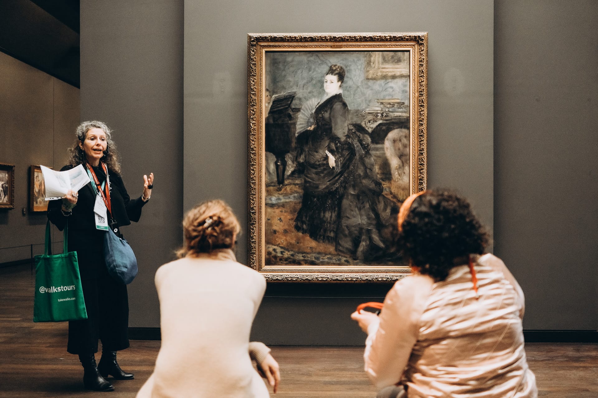 A tour guide explaining the details of a painting to a group of visitors at the Musée d'Orsay, standing in front of Portrait of Madame Charpentier by Pierre-Auguste Renoir.