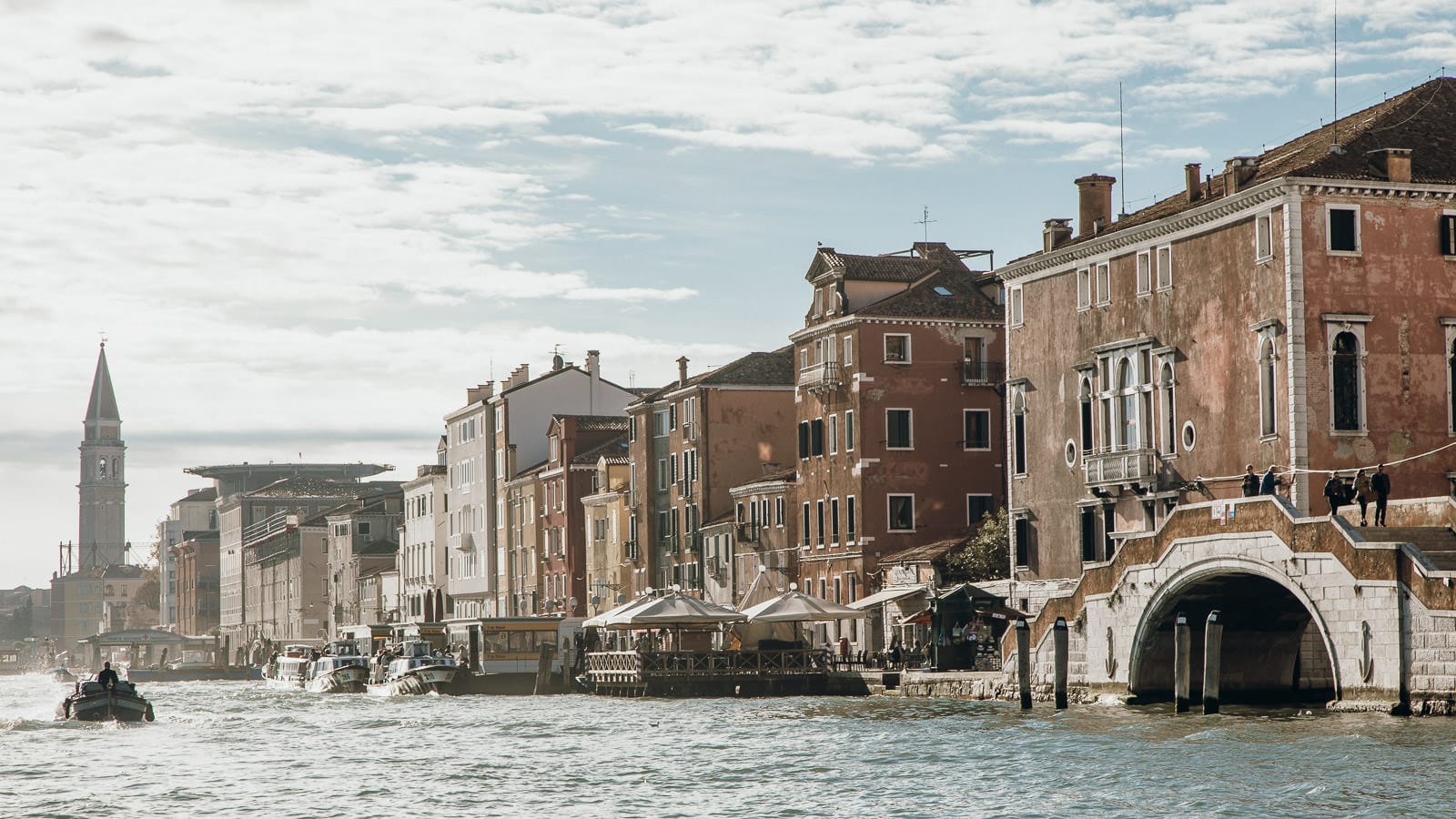 A scenic view of Venice’s Grand Canal with historic buildings, a bell tower in the distance, water taxis, and gondolas moving along the water under a partly cloudy sky.