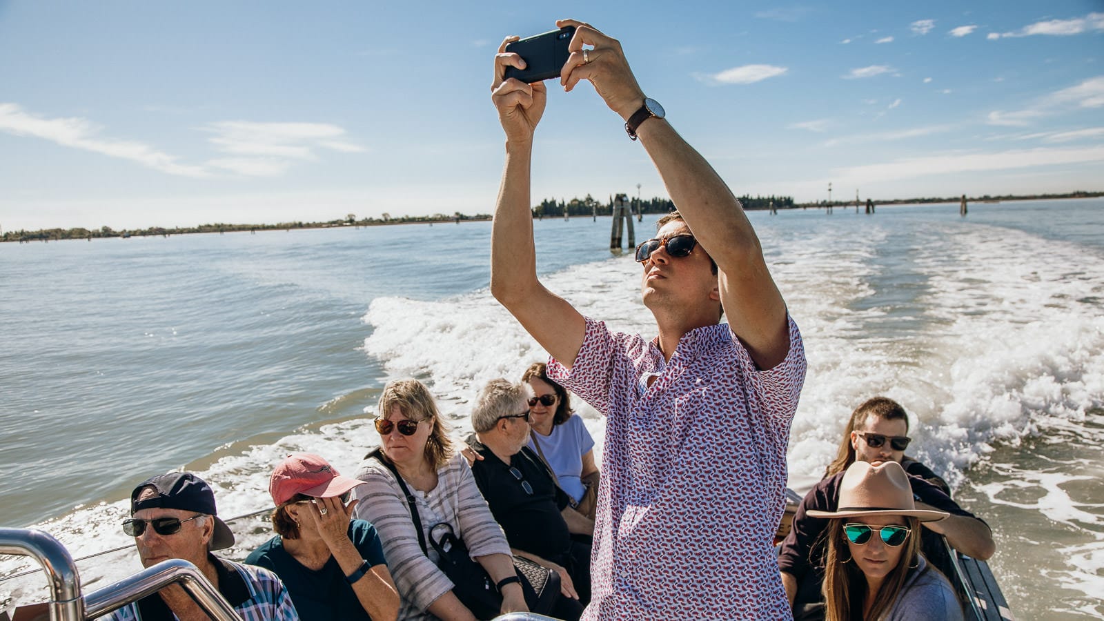 A group of people riding on a boat across calm waters on a sunny day. One man stands taking a photo with his phone while others sit, wearing sunglasses and hats, enjoying the ride to Murano and Burano from Venice. 