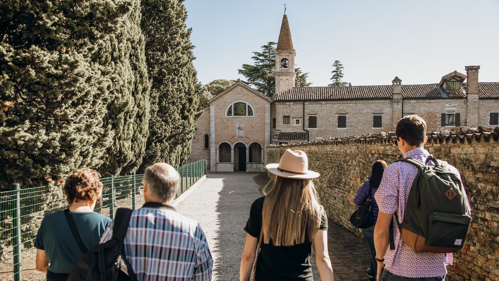 A group of five people walking down a stone pathway lined with tall cypress trees and old brick walls, heading toward a historic church with a bell tower and arched entrance.