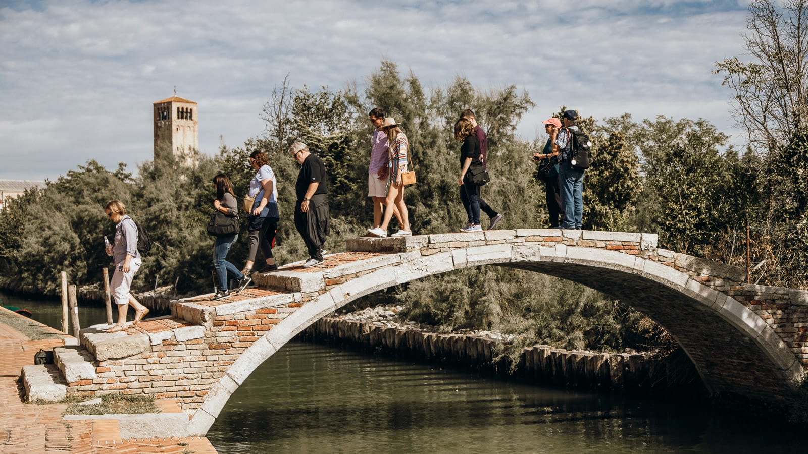 Tour group walking across a historic stone bridge on the Venetian lagoon near Murano and Burano, with a bell tower in the background.