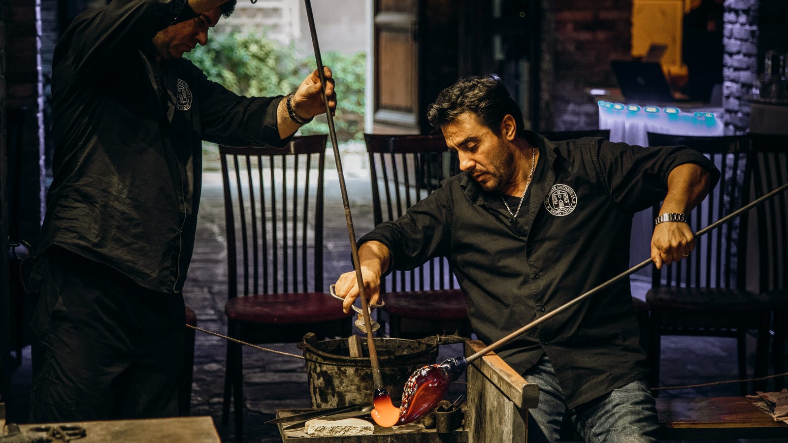 Two artisans carefully shaping molten glass using long metal rods during a glassblowing demonstration, with glowing orange glass being worked on in a workshop setting.
