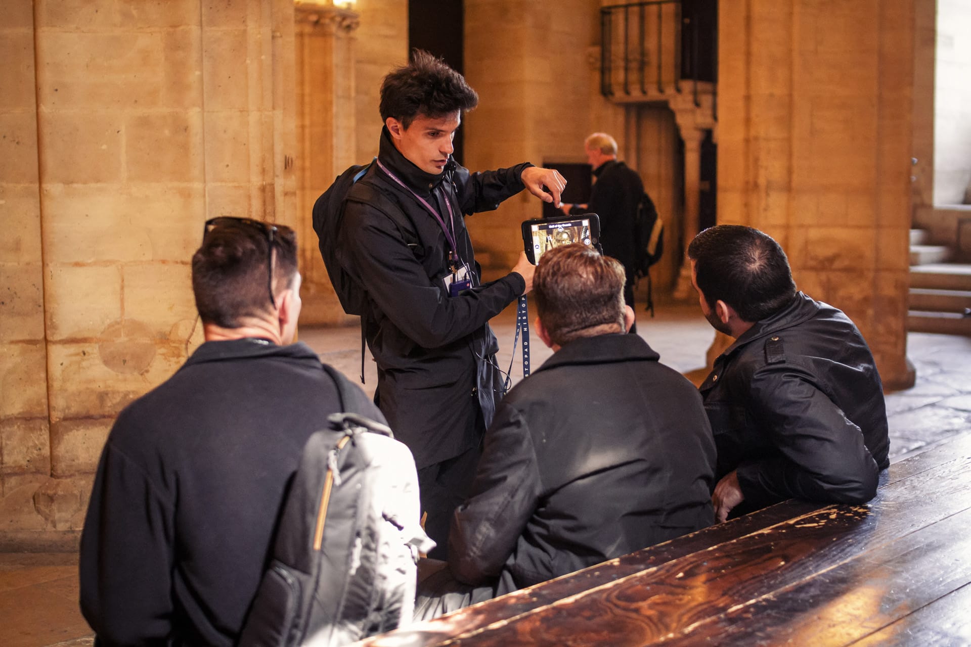  Tour guide showing a tablet with images to two seated visitors inside a historic stone building, engaging them in an educational explanation.