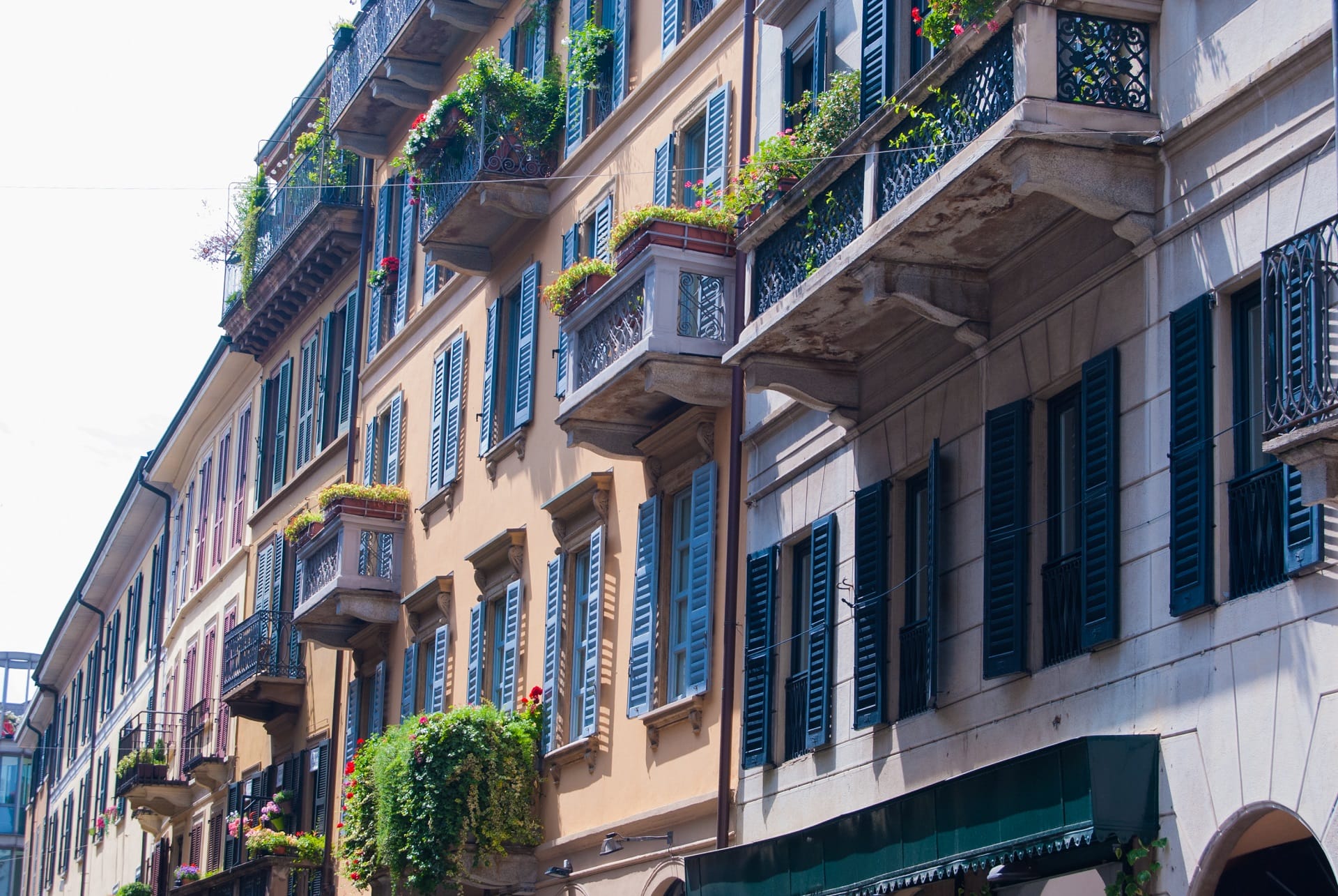 A street view of houses in the Brera area of Milan, with balconies and window boxes of greenery and red flowers.