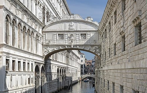 The Bridge of Sighs in Venice, Italy, arching gracefully over a narrow canal between the Doge’s Palace in Venice and the old prison, with a gondola passing in the background on the canal below.
