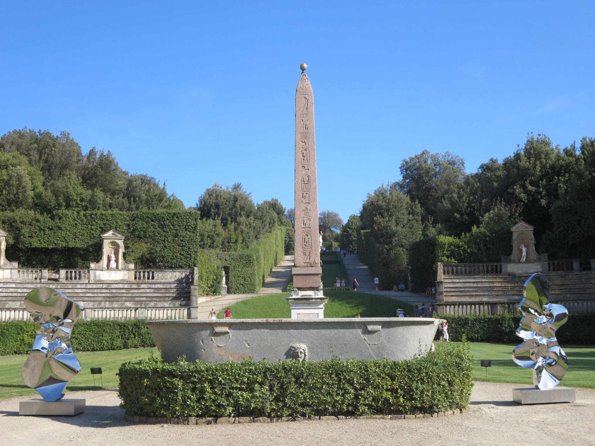 A 16 foot stone obelisk stands tall in front of ornate manicured hedges within the Boboli Gardens.