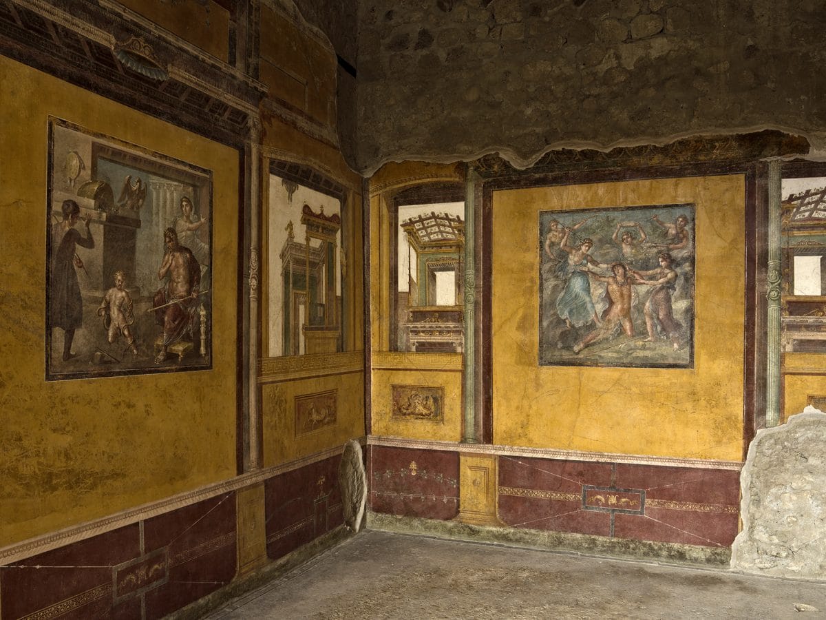 an internal view of the ancient wall murals inside the House of the Vettii in pompeii, in rich red and yellow tones with intricate scenes painted in central panels.