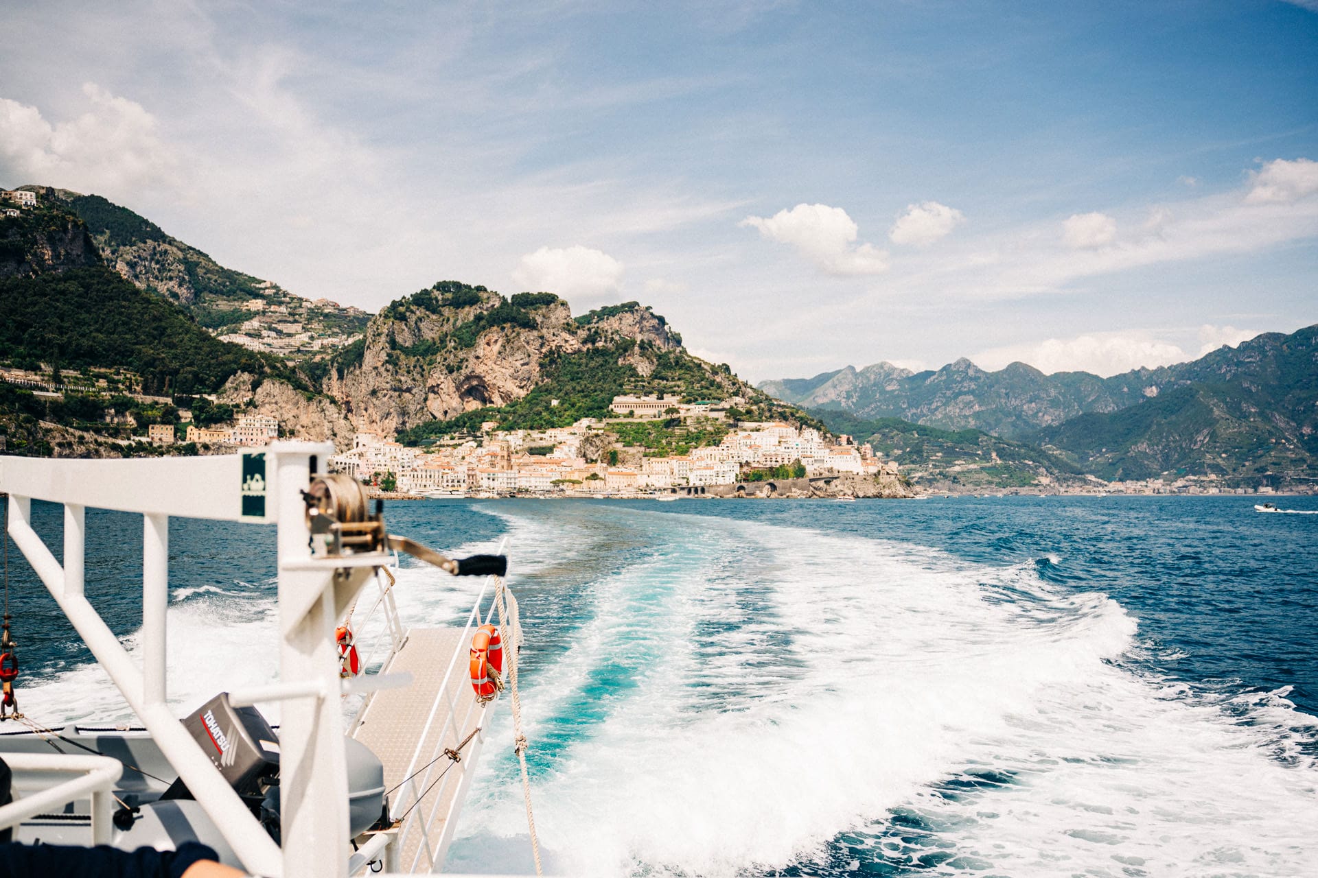 A panoramic view of one of the villages the amalfi coast, flanked by rocky cliffs and mountainous terrain, from the deck of a boat out at sea.