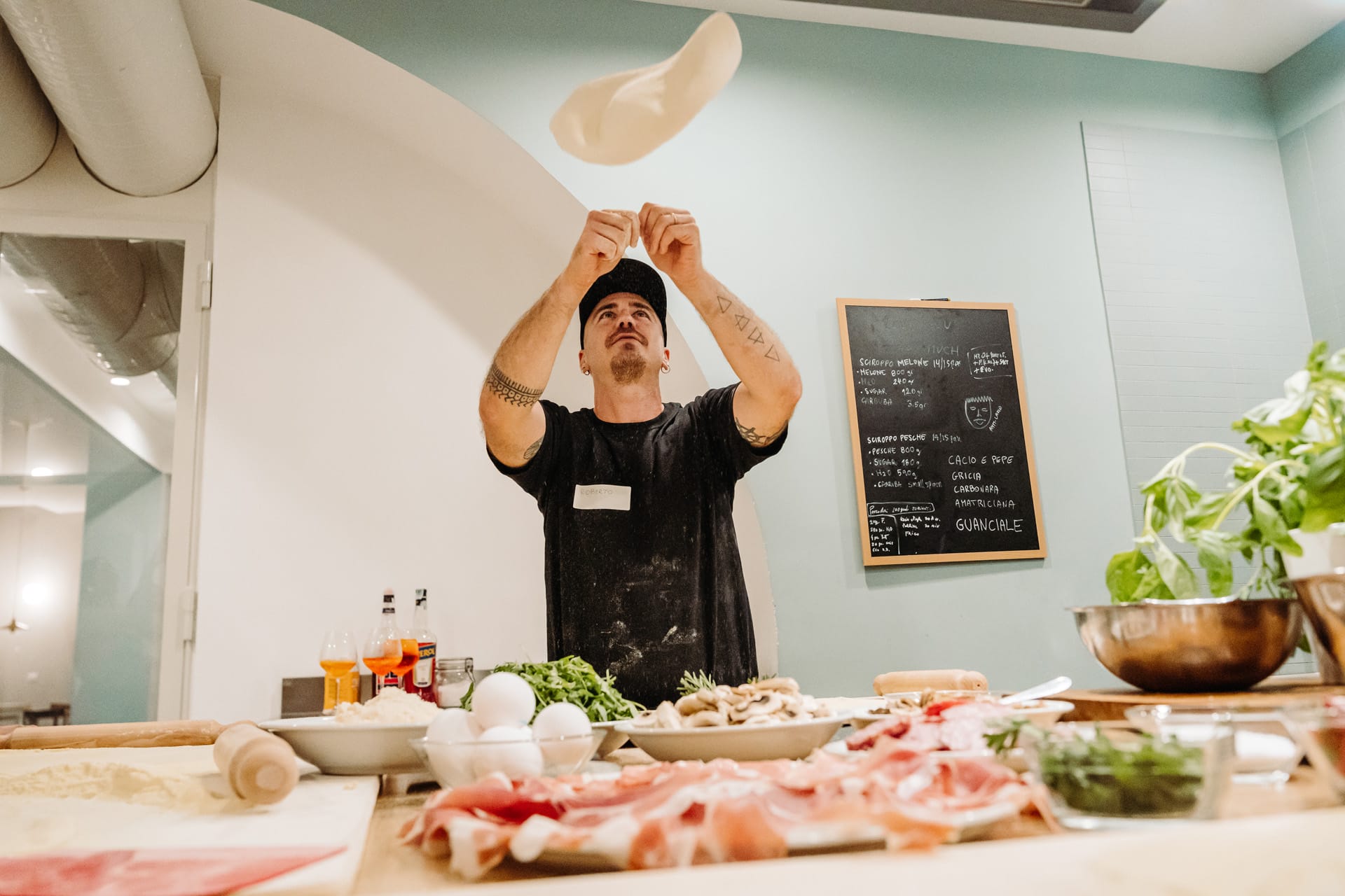 A man ina black tshirt and cap stands behind a wooden counter laden with fresh food, meat, and pizza toppings, as he throws the dough in the air to stretch it 