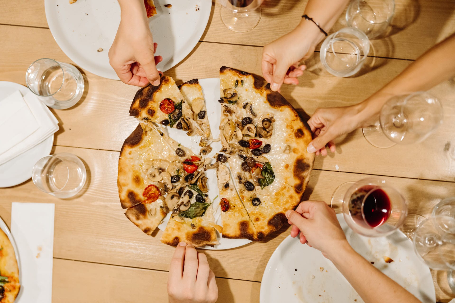 A freshly made Italian thin crust pizza with traditional toppings of tomato, cheese and olives in the centre of a table with 5 hands reaching in to take a slice 