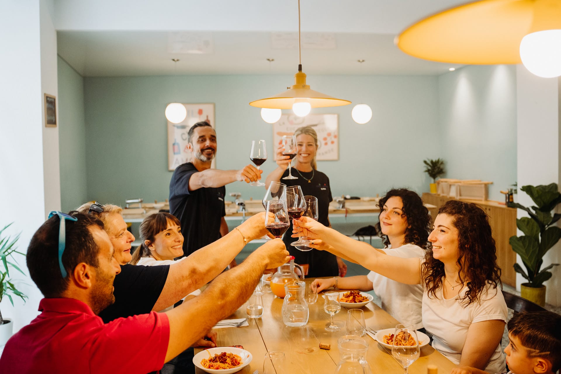 A group of 6 people sitting at a big wooden table with plates of pasta, and 2 chefs standing at the head of the table,  toast in the centre of the photo with wine glasses 