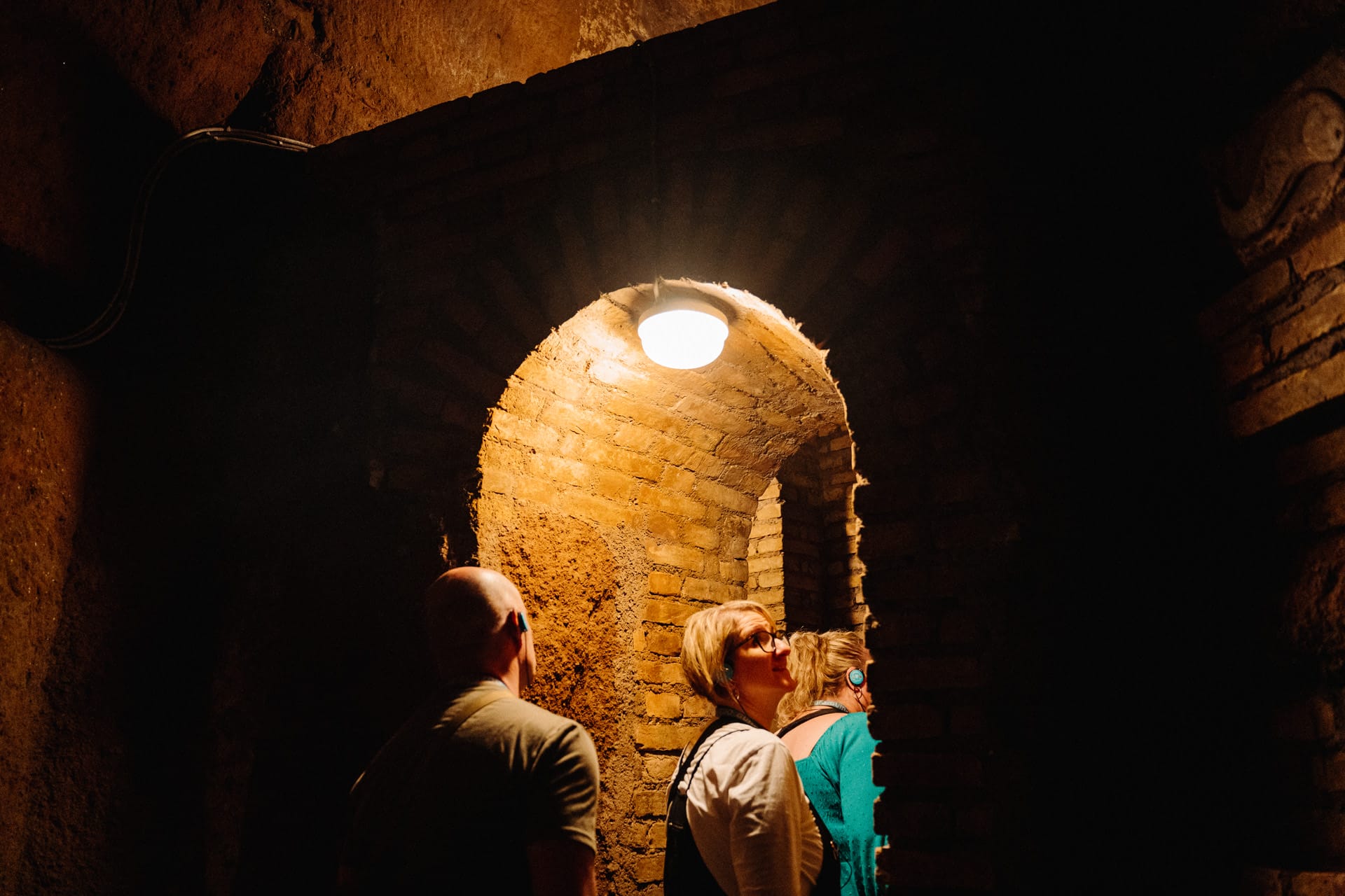 A small group of visitors walk through an archway that has been lit up, in the middle of an underground tunnel within the Rome catacombs