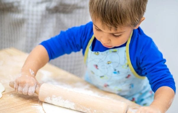A young boy on a cooking in Rome for Kids. He is wearing a blue jumper and multicolour apron, with a rolling pin covered in flour, rolling out pizza dough on a wodden counter top 