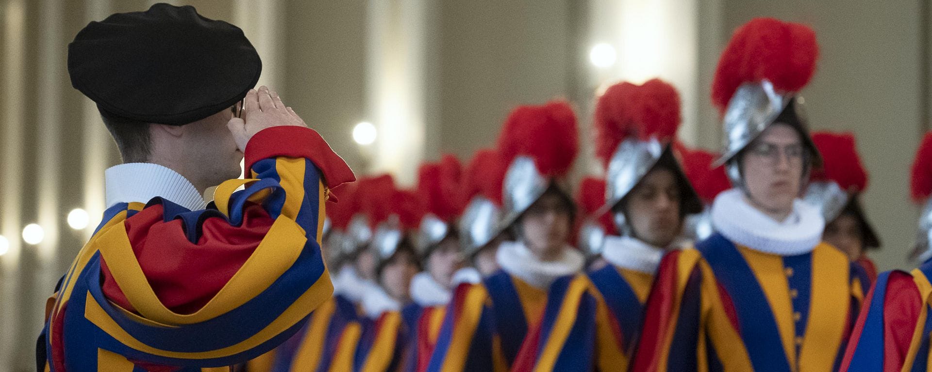 A member of the swiss guard in a black beret and red white and blue uniform salutes to a troop who also wear ruffles neck collar and silver armoured helmet with red feather plumes
